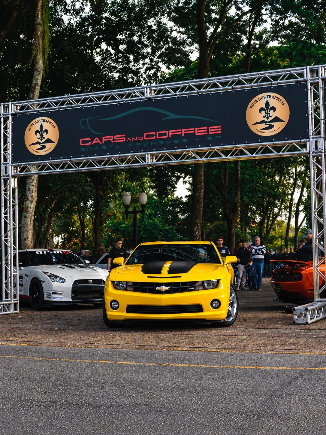 Camaro amarelo passando sob o pórtico do Cars and Coffee Tremembé, com GT-R branco ao lado e público ao fundo